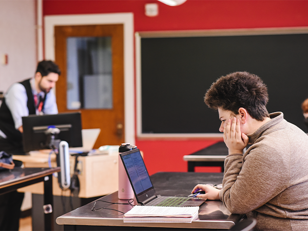 A student, head in hands, works on a laptop in a classroom. An instructor stands at a desk nearby.